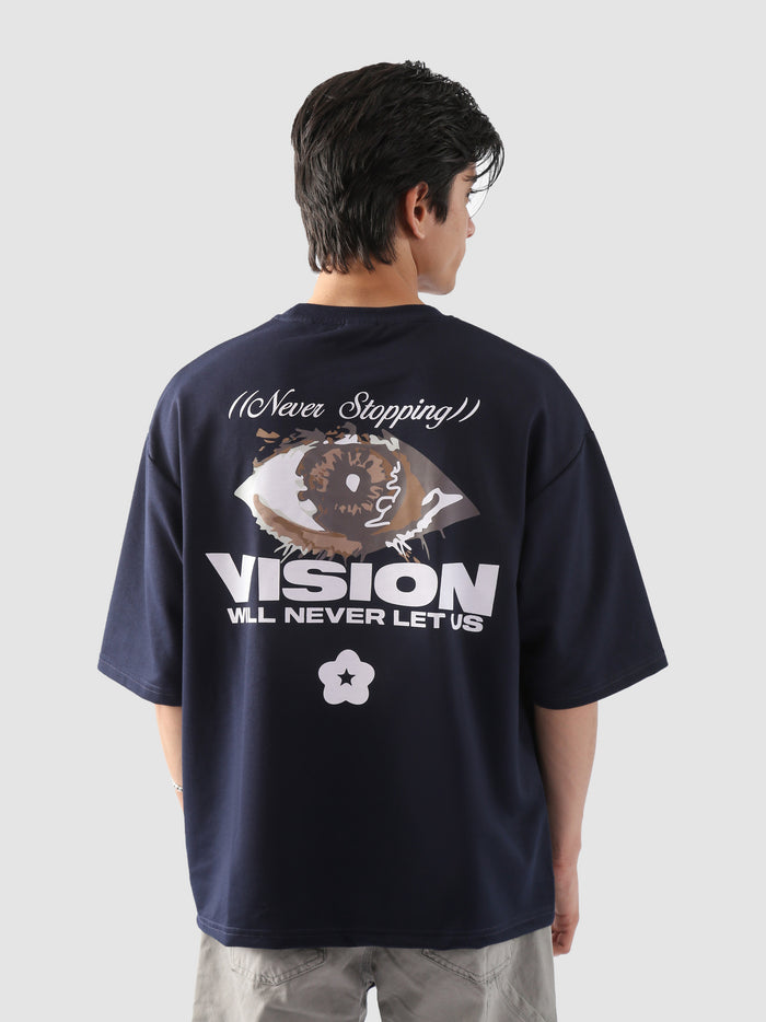 Close-up back view of a male model wearing the navy "Never Stopping Culture" t-shirt, featuring a bold eye graphic and the phrase “VISION WILL NEVER LET US” in puff and screen print – symbolizing unwavering drive and hustle.