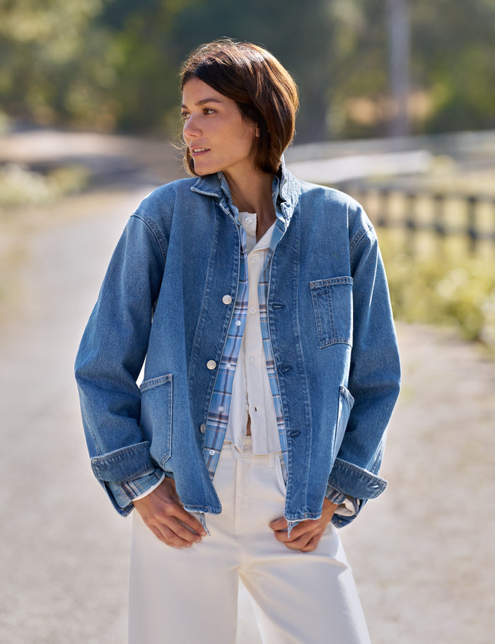 A woman wearing a light blue denim jacket over a blue plaid shirt and white pants, standing outdoors on a sunlit path with a blurred natural background.