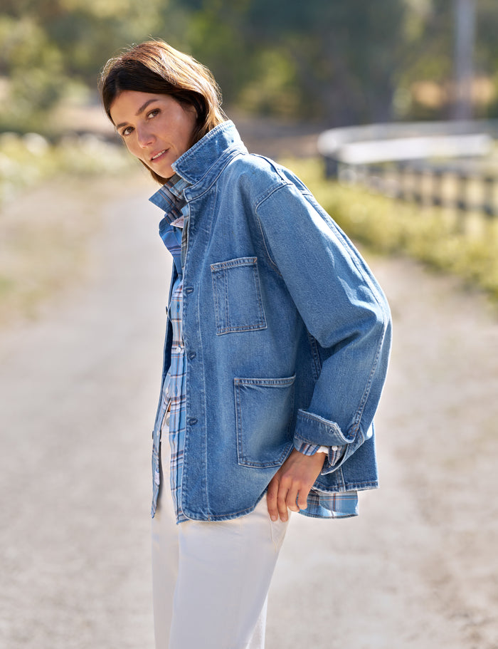 A woman wearing a light blue denim jacket over a plaid shirt and white pants, standing on a sunlit dirt path outdoors.