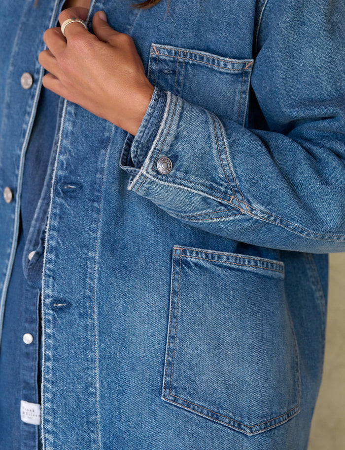 Close-up of a person wearing a medium-wash blue denim button-up shirt jacket with front pockets and a visible hand adjusting the collar.