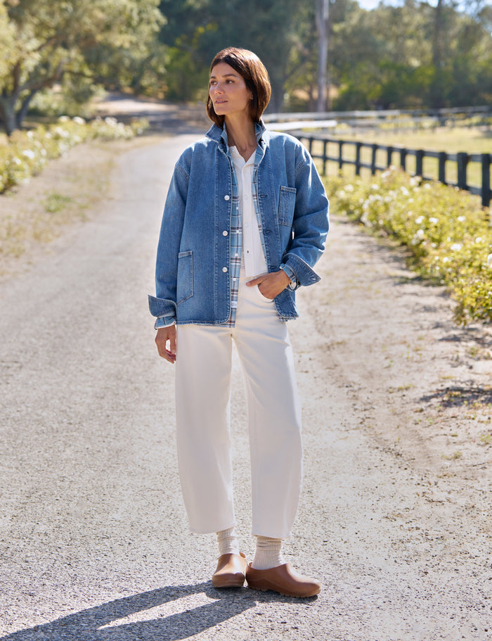 A woman stands on a sunlit gravel path outdoors wearing a light blue denim jacket over a white top and white wide-leg pants, with tan clogs and a watch.