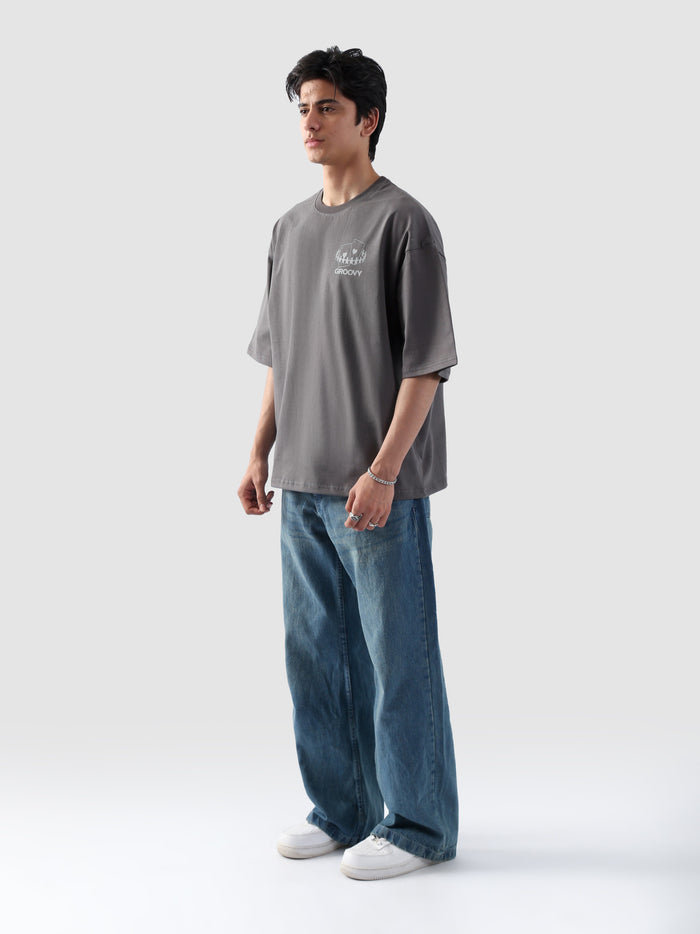 Male model wearing stone grey oversized t-shirt in Pakistan by Groovy – drop shoulder, baggy fit graphic tee styled with loose denim jeans