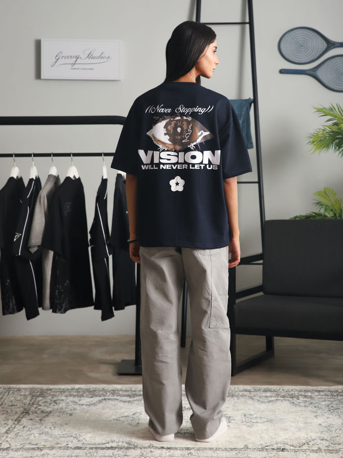 Back view of a woman wearing the "Never Stopping Culture" navy blue graphic t-shirt featuring an eye design and bold text, styled with light grey cargo pants in a minimalist studio setup.