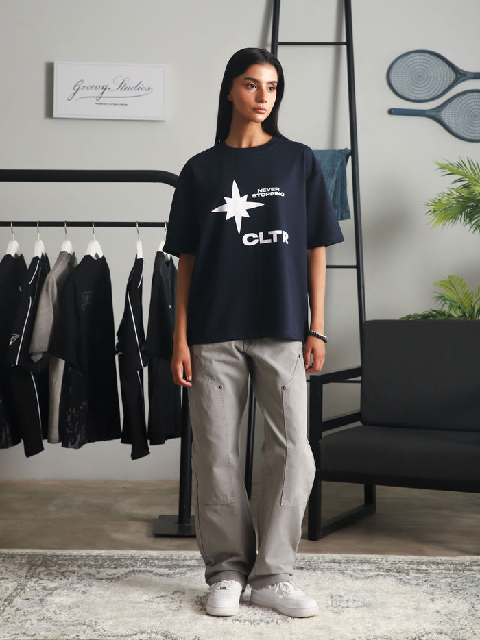 Front view of a woman wearing the navy "Never Stopping Culture" t-shirt with bold white star and CLTR logo, styled with grey cargo pants and white sneakers, standing in a minimal studio setup with clothing racks and indoor plants.