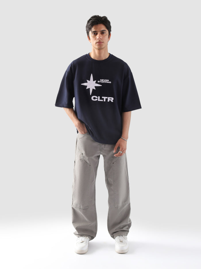 Front-facing shot of a male model wearing the "Never Stopping Culture" navy blue tee with bold white CLTR star graphic, paired with light grey baggy cargo pants and white sneakers, set against a minimalist grey backdrop.